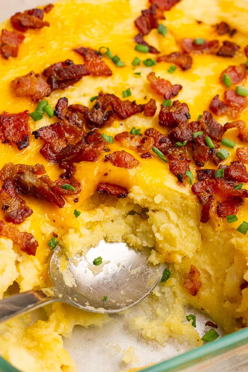 A spoon scooping into the soft, cheesy potatoes underneath a layer of melted cheddar, bacon, and chives.