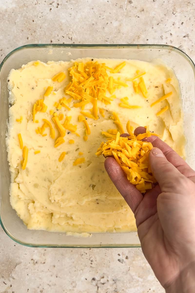 A hand sprinkling shredded cheddar cheese over the mashed potato mixture in a baking dish.