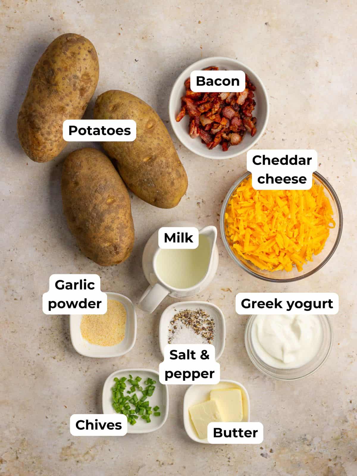A flat lay of all the ingredients for twice baked mashed potatoes including potatoes, cheese, bacon, milk, butter, Greek yogurt, garlic powder, and chives.