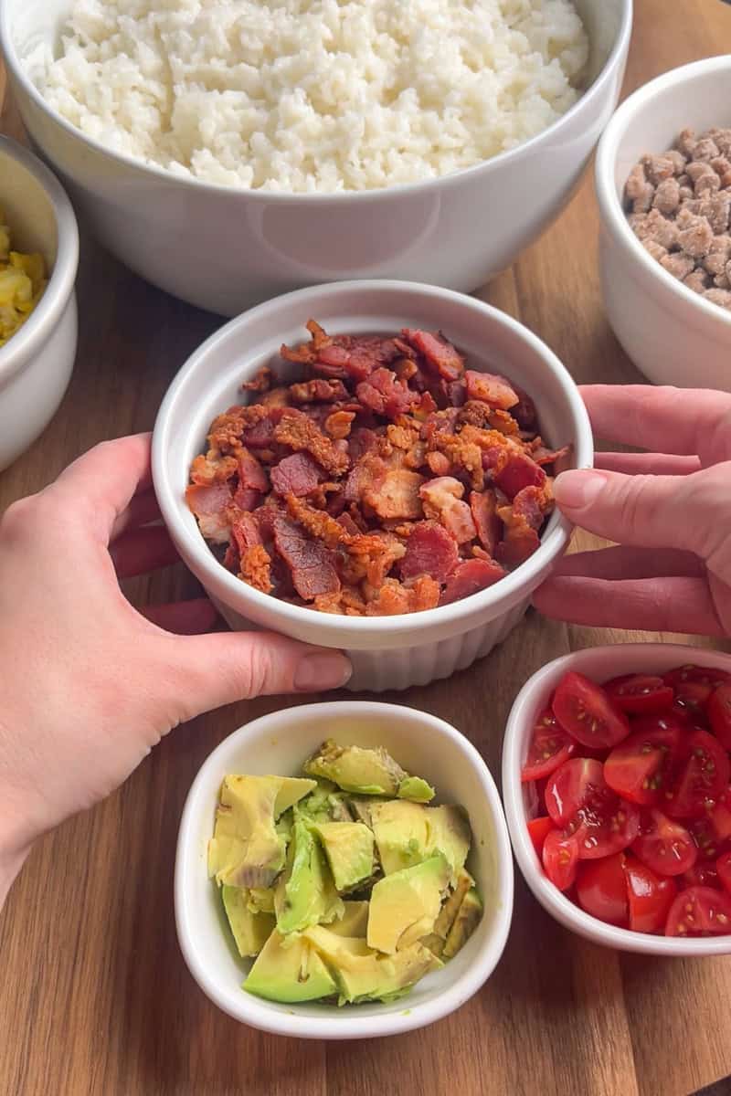 Hands holding a bowl of crispy bacon with other breakfast rice bowl ingredients in the background.