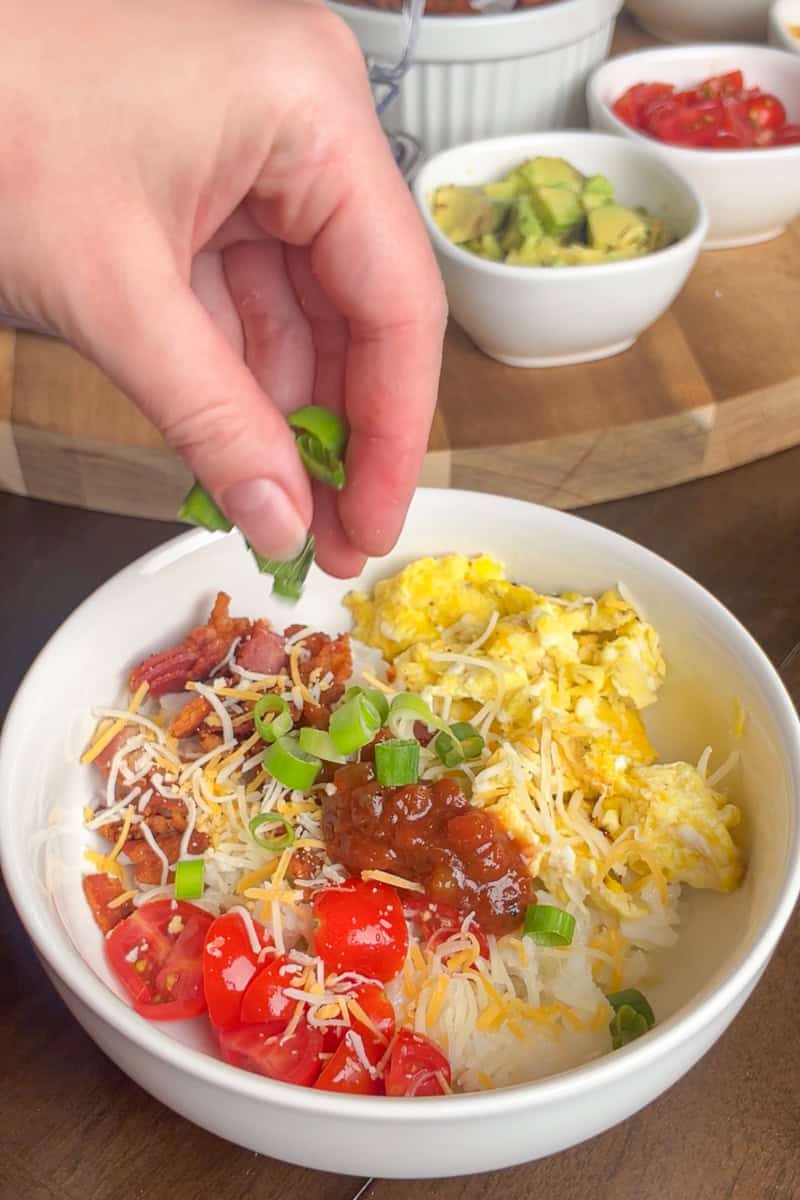A hand sprinkling green onions over a breakfast rice bowl with eggs, bacon, cheese, and tomatoes.