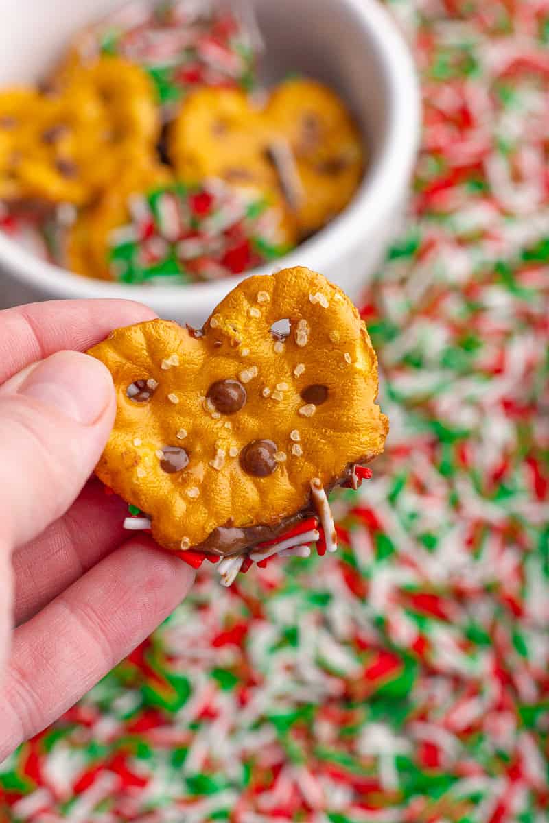 A hand holds a single pretzel dipped in chocolate and caramel with sprinkles in the background.