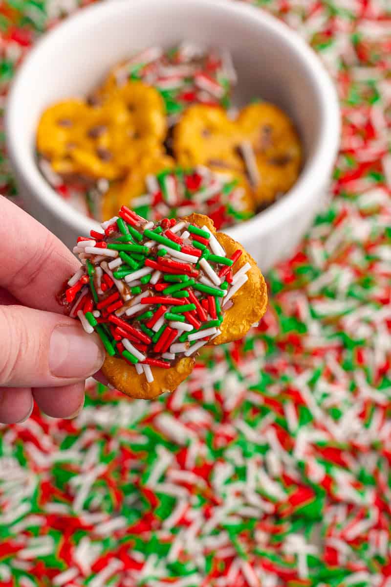 A close-up of a christmas crack pretzel covered in red, green, and white sprinkles held in front of a small bowl.