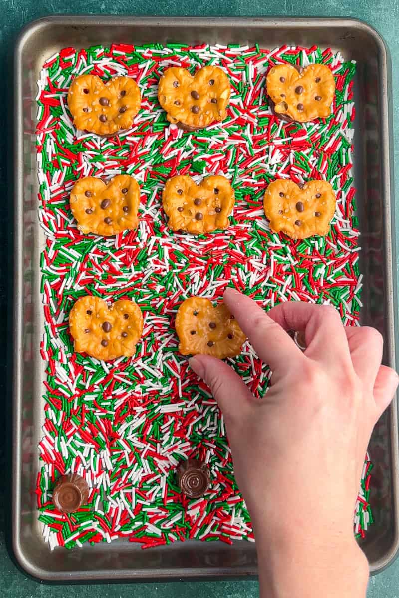 A hand presses a pretzel into a softened Rolo candy on a sprinkle-covered baking sheet.
