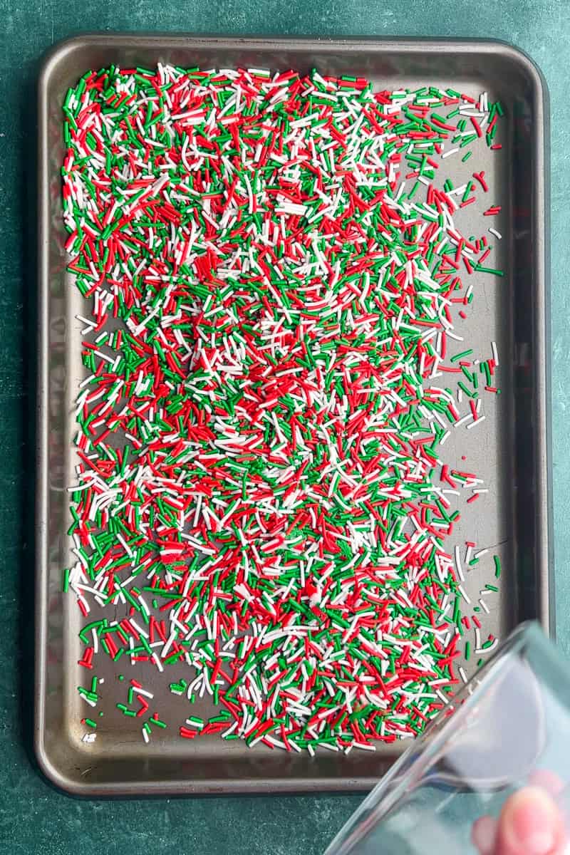 A baking sheet is covered with red, green, and white sprinkles before adding pretzels and Rolos.