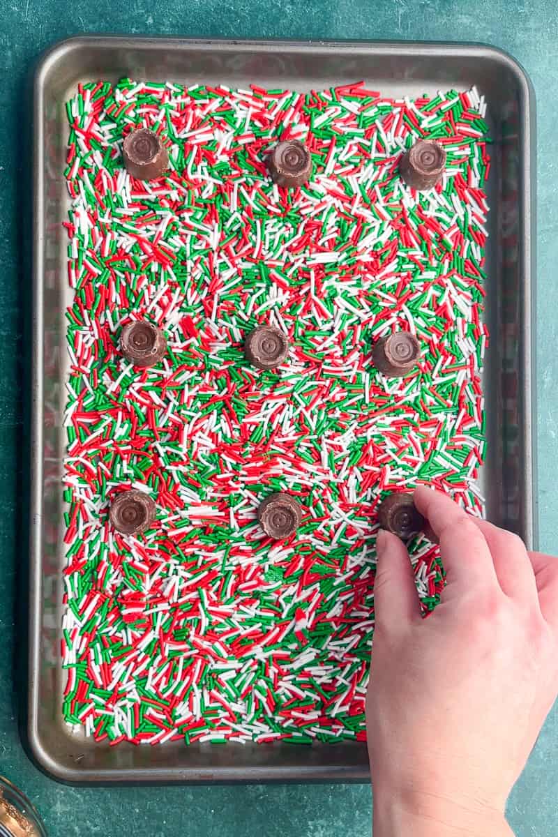 Unwrapped Rolo candies are spaced evenly on a baking sheet layered with Christmas sprinkles.
