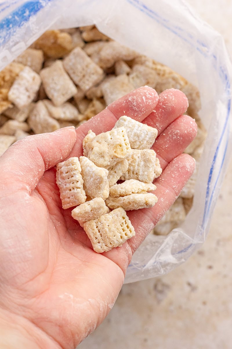 A hand holding cookie butter puppy chow pieces dusted with powdered sugar over an open bag.