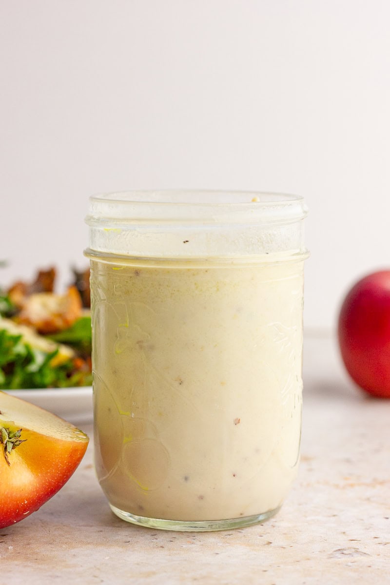 A glass mason jar filled with creamy apple cider vinaigrette sits on a countertop with sliced apples nearby.