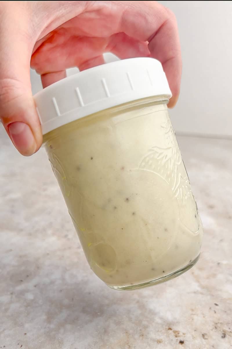 A hand holds a sealed mason jar of creamy apple cider vinaigrette while tilting it slightly.