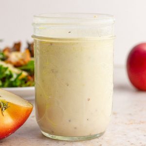 A mason jar of creamy apple cider vinaigrette is displayed on a countertop with sliced apples and salad in the background.