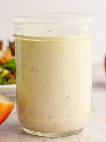 A mason jar of creamy apple cider vinaigrette is displayed on a countertop with sliced apples and salad in the background.
