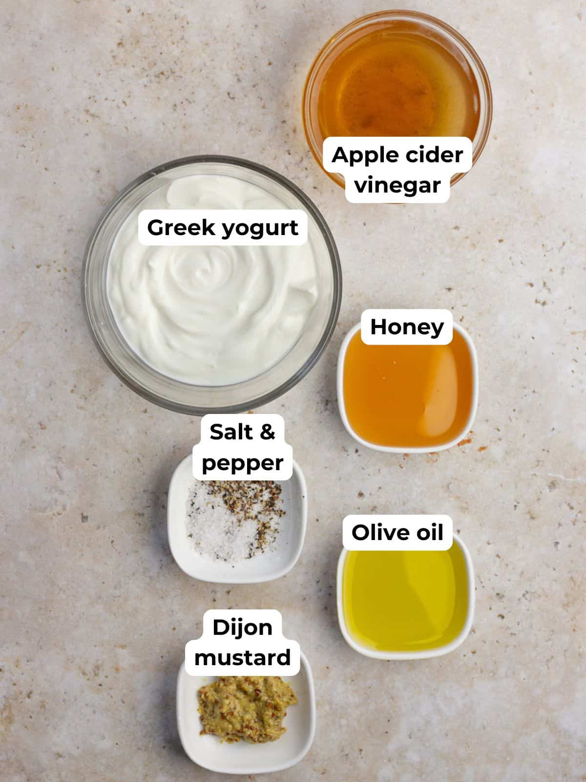 Bowls of Greek yogurt, apple cider vinegar, honey, olive oil, Dijon mustard, salt, and pepper are arranged on a countertop.