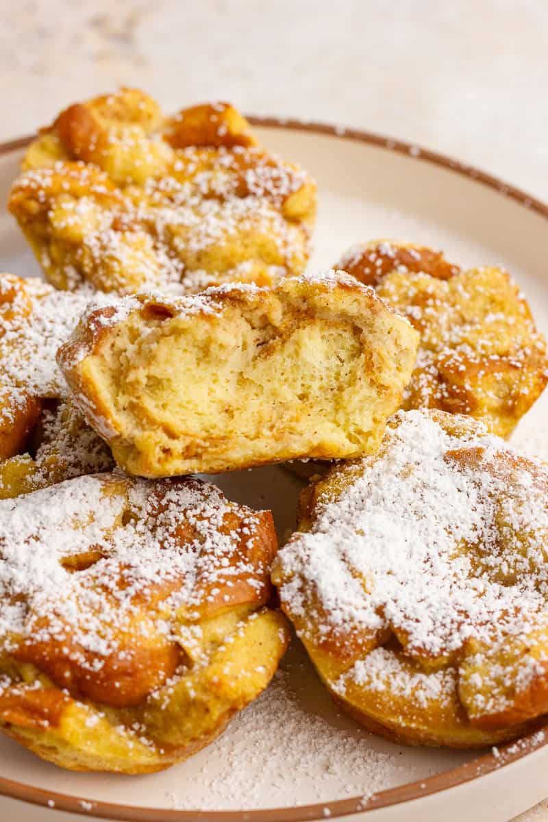 A plate of golden french toast cups topped with powdered sugar, with one cup cut open to show the soft interior.