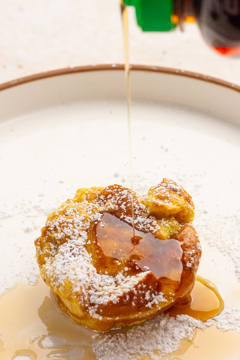 A french toast cup dusted with powdered sugar while maple syrup is being poured over the top.