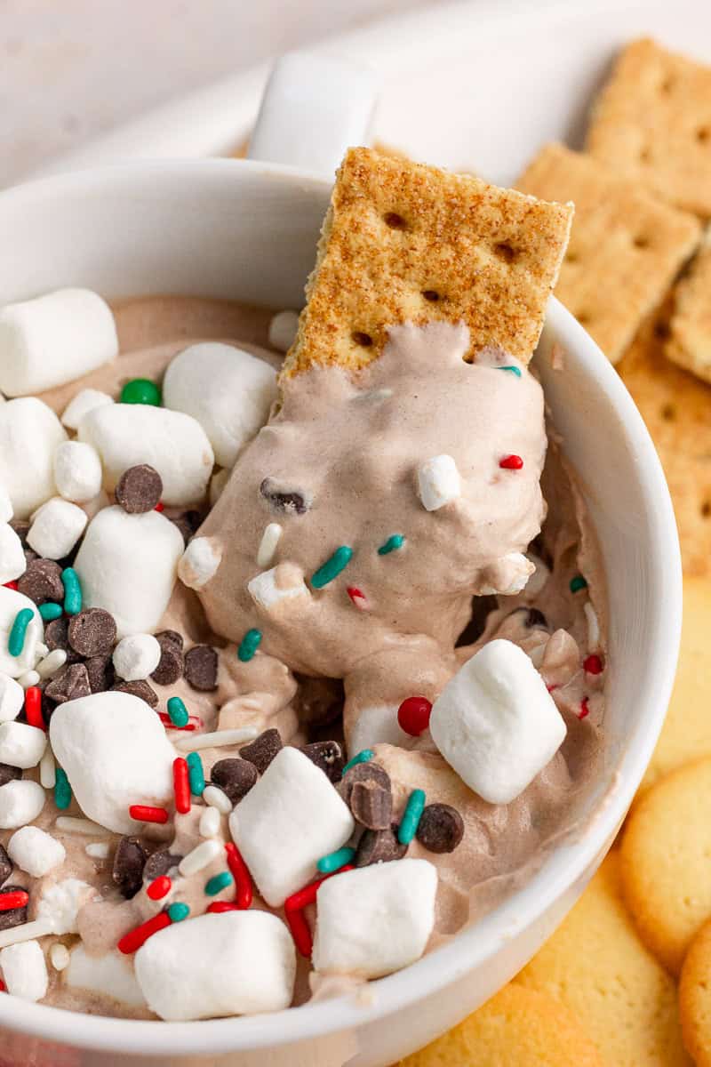 A graham cracker dipped into creamy hot cocoa dip topped with marshmallows and festive sprinkles.