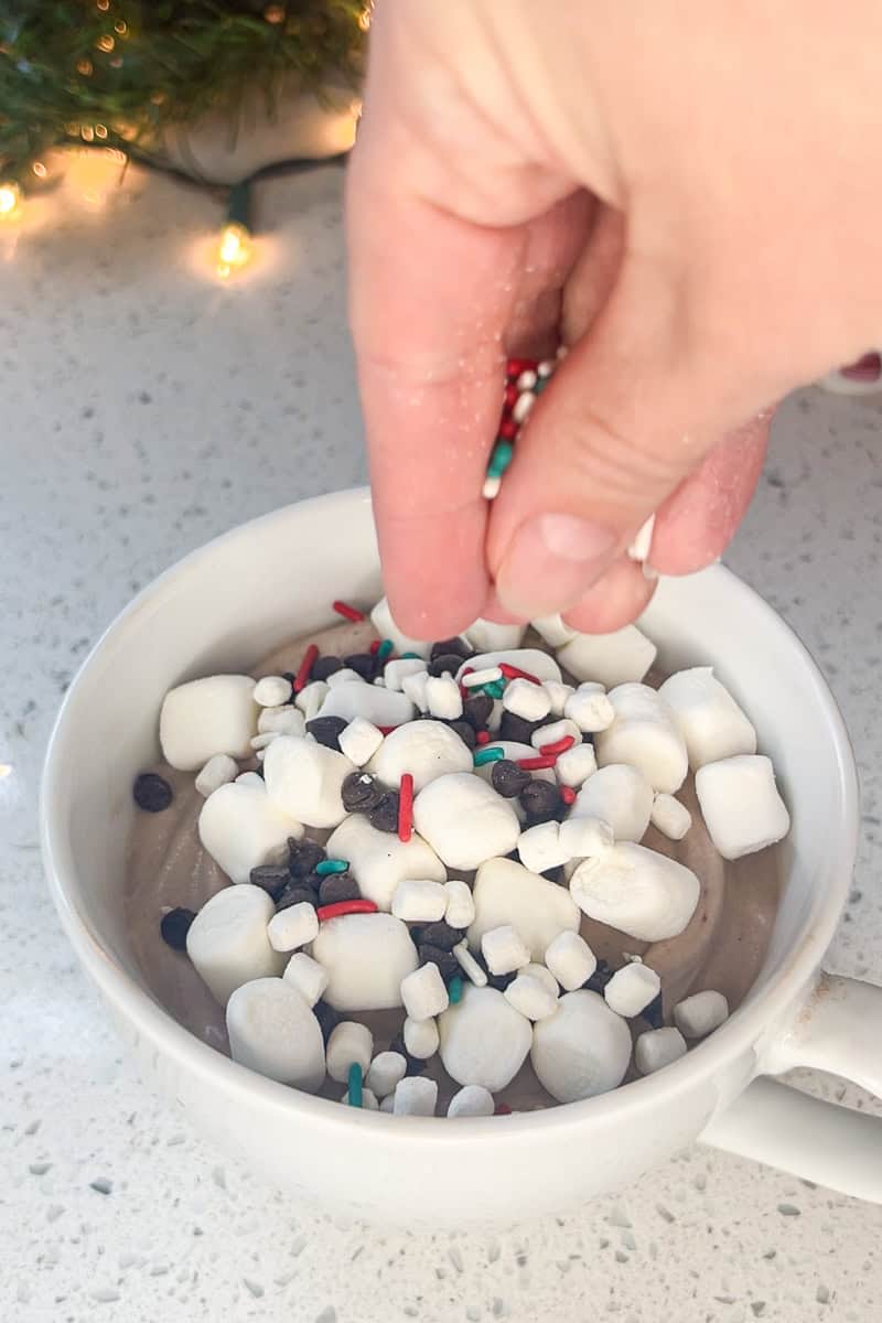 Hand sprinkling marshmallows and sprinkles over a bowl of hot cocoa dip on a holiday countertop.