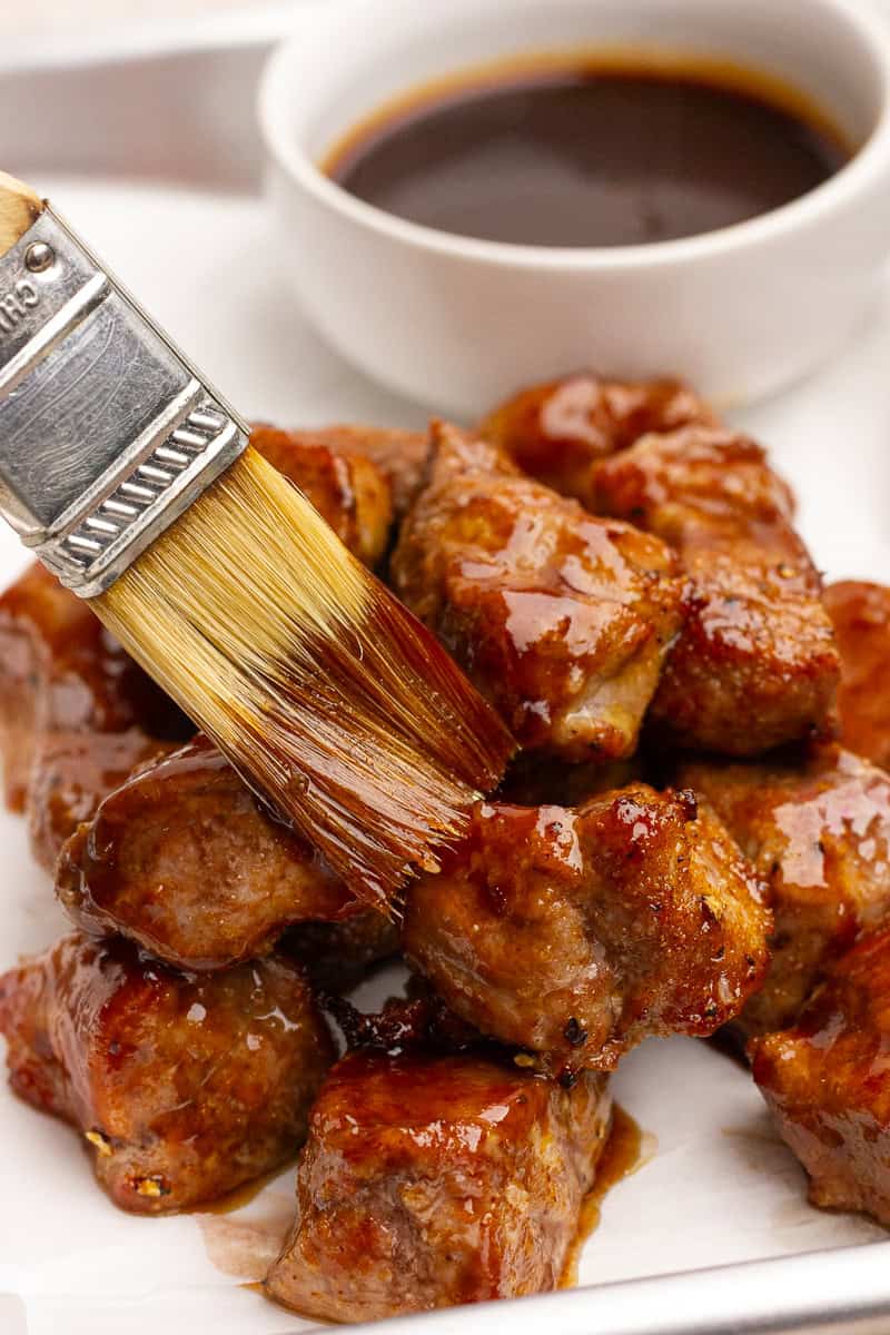 A brush is coating the hot pork bites with a glossy layer of BBQ sauce.