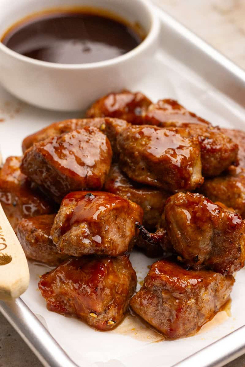 A pile of saucy pork tenderloin bites sits on a tray with a small bowl of BBQ sauce in the background.