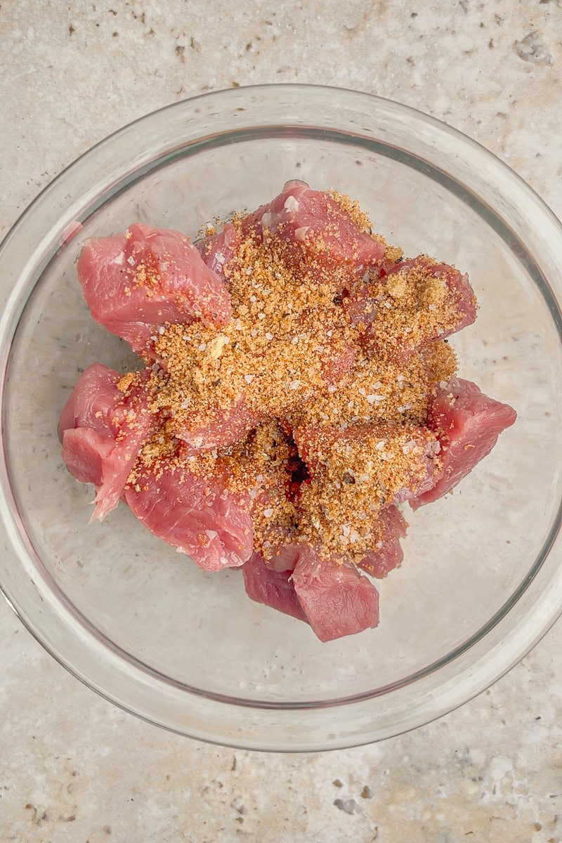 Raw pork tenderloin chunks sit in a glass bowl topped with dry BBQ seasoning.