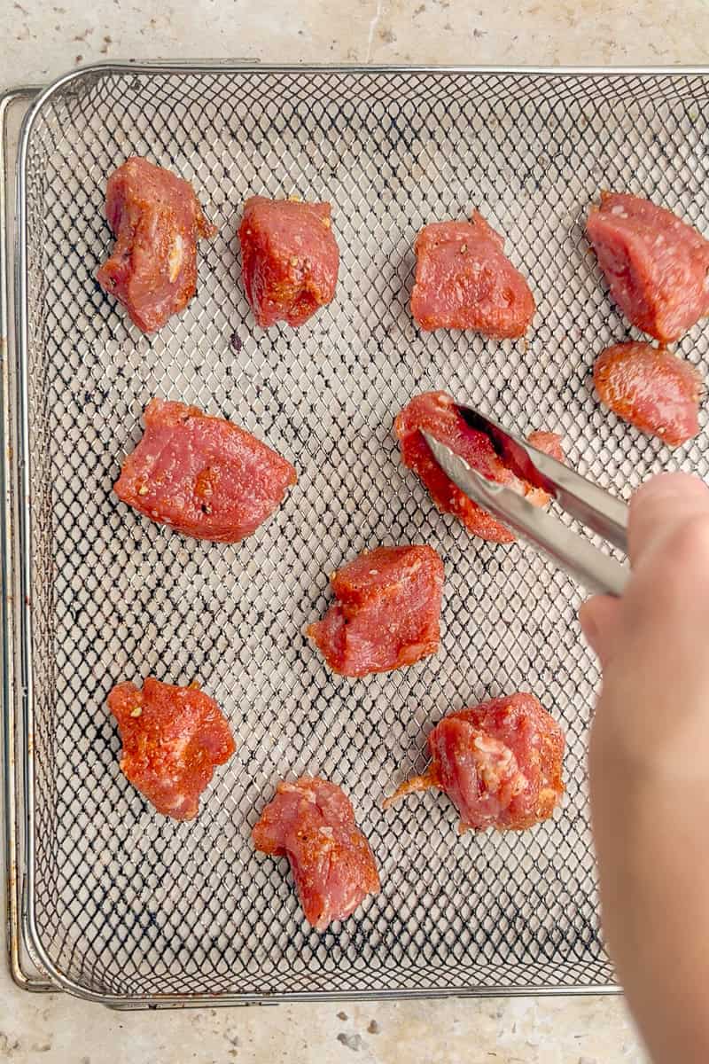 Seasoned pork tenderloin chunks are arranged on an air fryer rack while a hand uses tongs to place one piece down.
