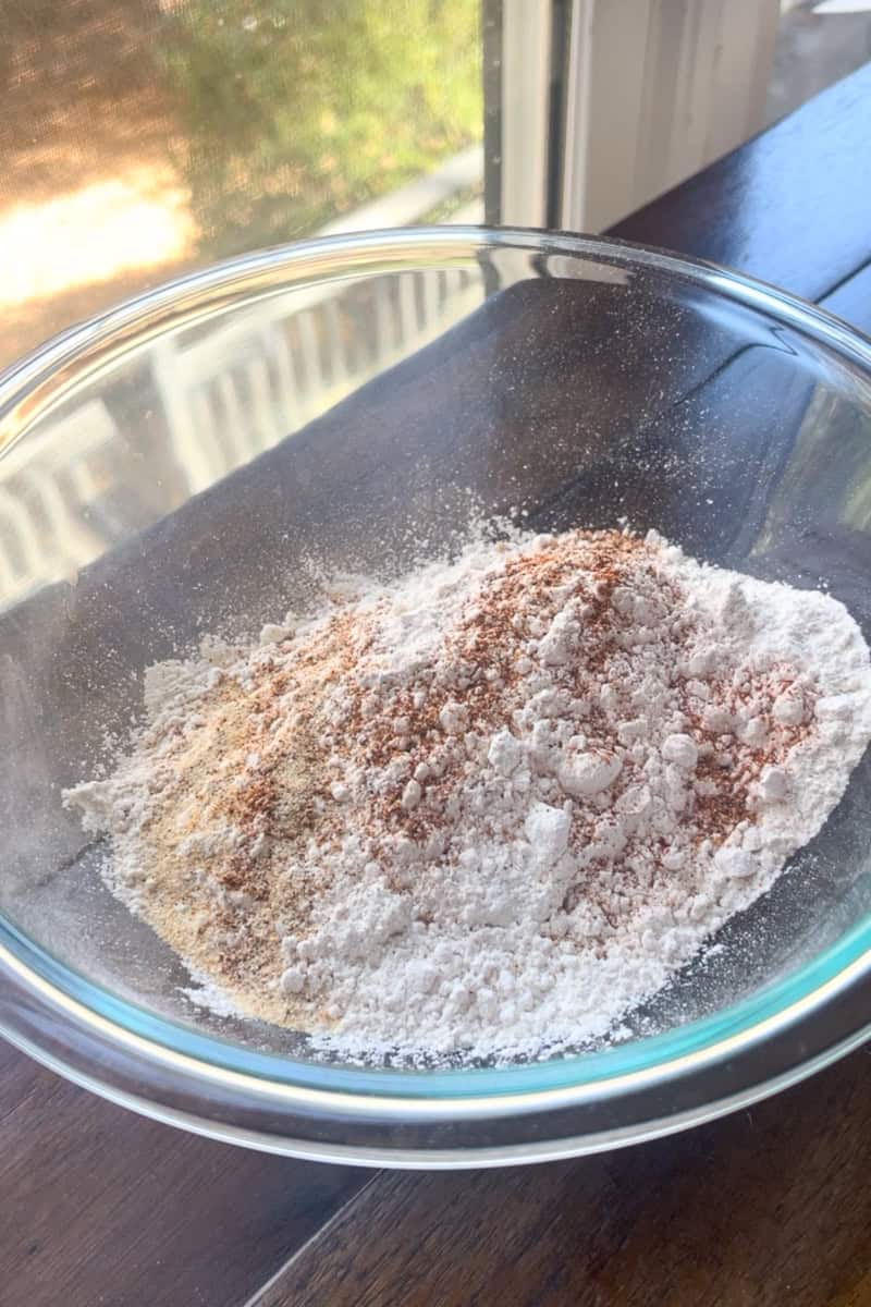 A bowl of the dry flour mixture with seasonings sits on a wooden surface near a window.