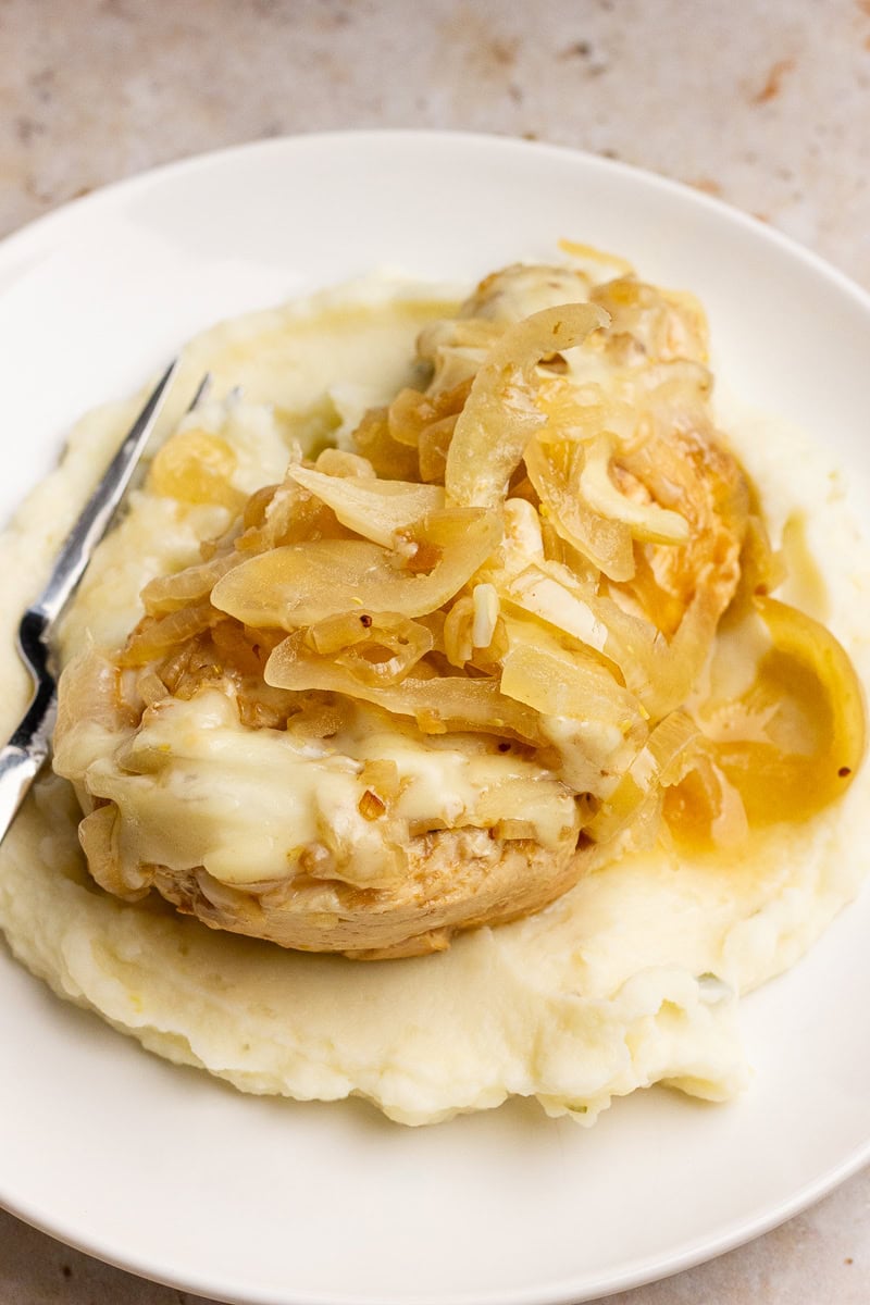 A chicken breast topped with soft cooked onions is served over mashed potatoes on a white plate.