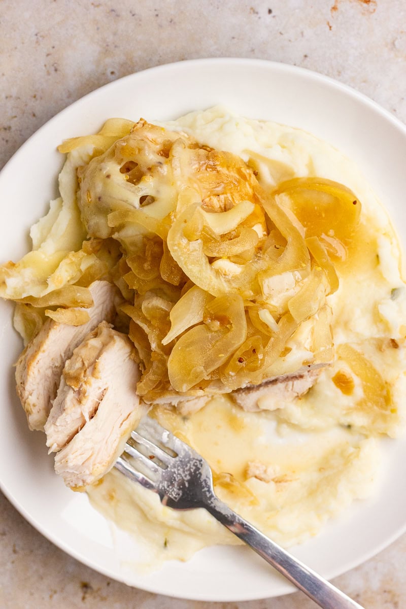 A plate of sliced French onion chicken sits over mashed potatoes with onions and broth on top.