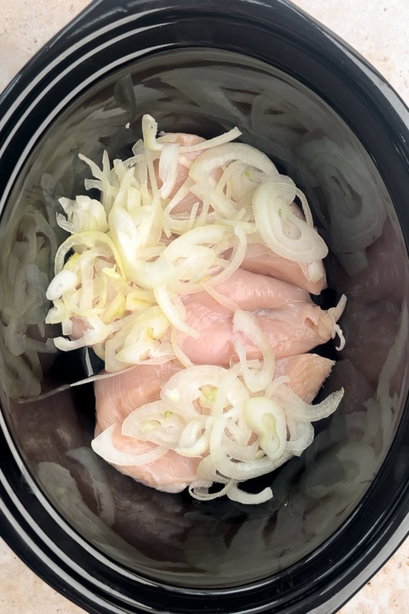 Raw chicken breasts are placed in a slow cooker and topped with thinly sliced onions.