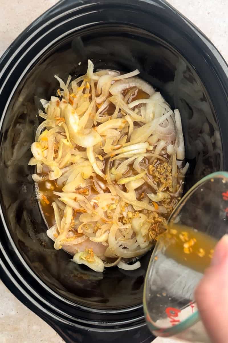 Broth and seasonings are being poured over chicken and onions in a slow cooker.