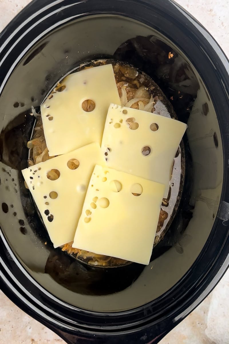 Four slices of Swiss cheese are layered on top of the cooked chicken and onions in a slow cooker.
