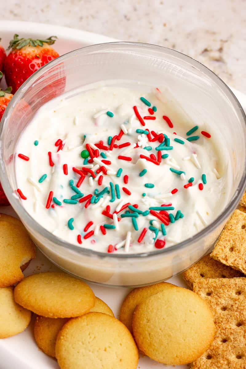 A close-up of sugar cookie dip in a glass bowl with festive sprinkles and cookies arranged around the plate.