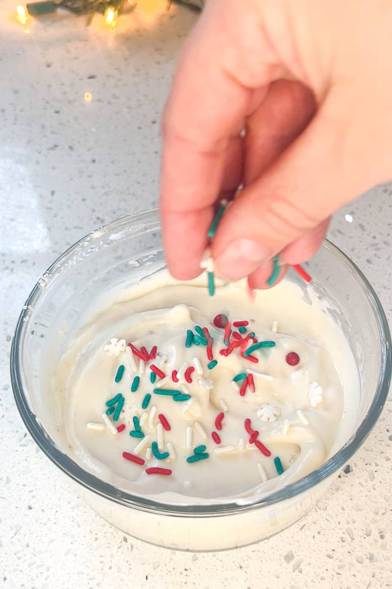A hand sprinkling red, green, and white sprinkles over a bowl of sugar cookie dip.