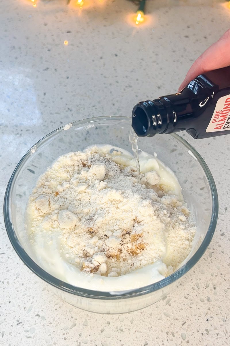 Sugar cookie mix and almond extract being added to vanilla yogurt in a mixing bowl.