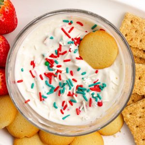A bowl of creamy sugar cookie dip topped with red, green, and white sprinkles, surrounded by vanilla wafers, graham crackers, and strawberries.