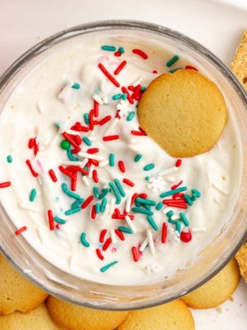 A bowl of creamy sugar cookie dip topped with red, green, and white sprinkles, surrounded by vanilla wafers, graham crackers, and strawberries.