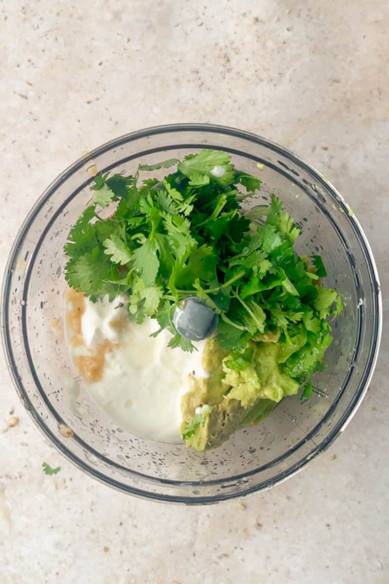 Ingredients for avocado lime crema including avocado, Greek yogurt, lime juice, garlic, and cilantro in a food processor.