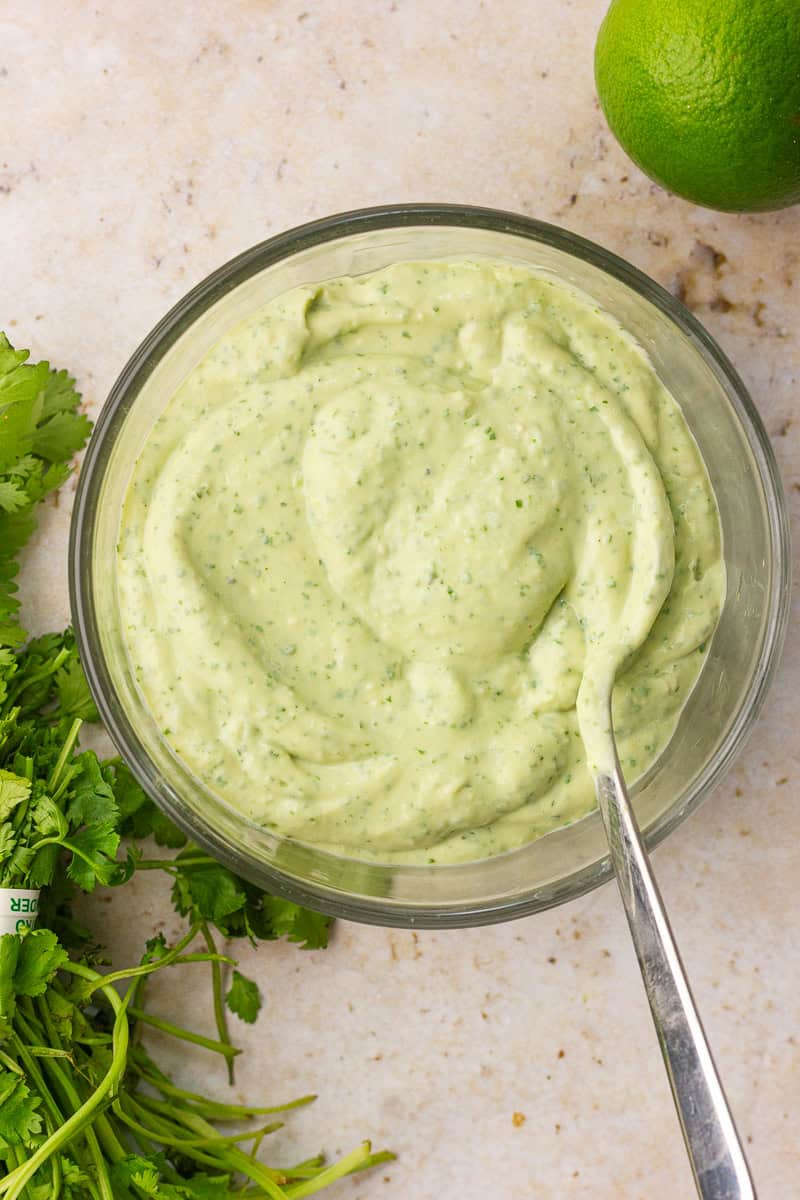 A bowl of creamy avocado lime crema with a spoon, surrounded by fresh cilantro and a whole lime.
