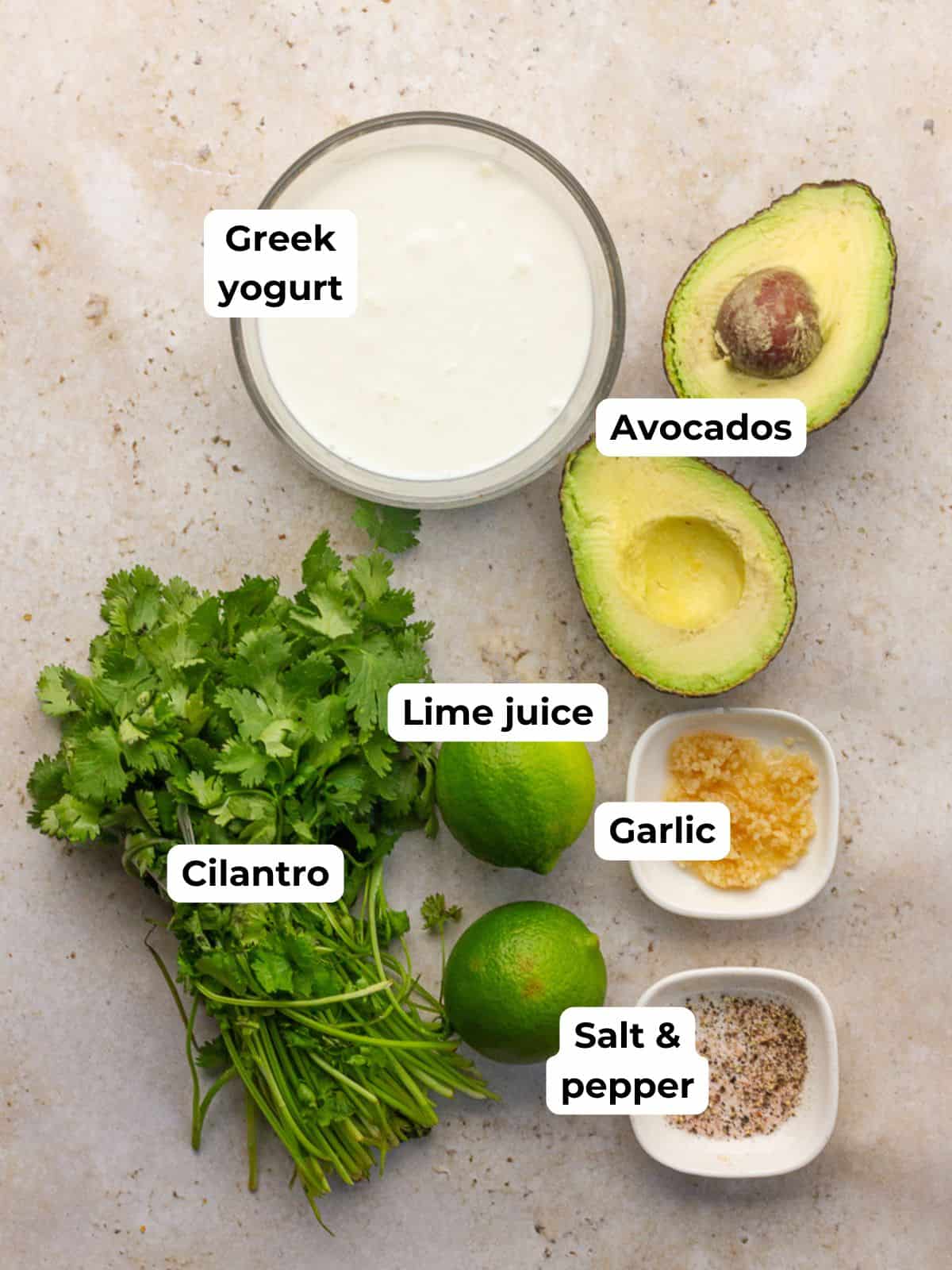 Ingredients for avocado lime crema including Greek yogurt, avocados, lime juice, garlic, cilantro, and salt and pepper laid out on a surface.