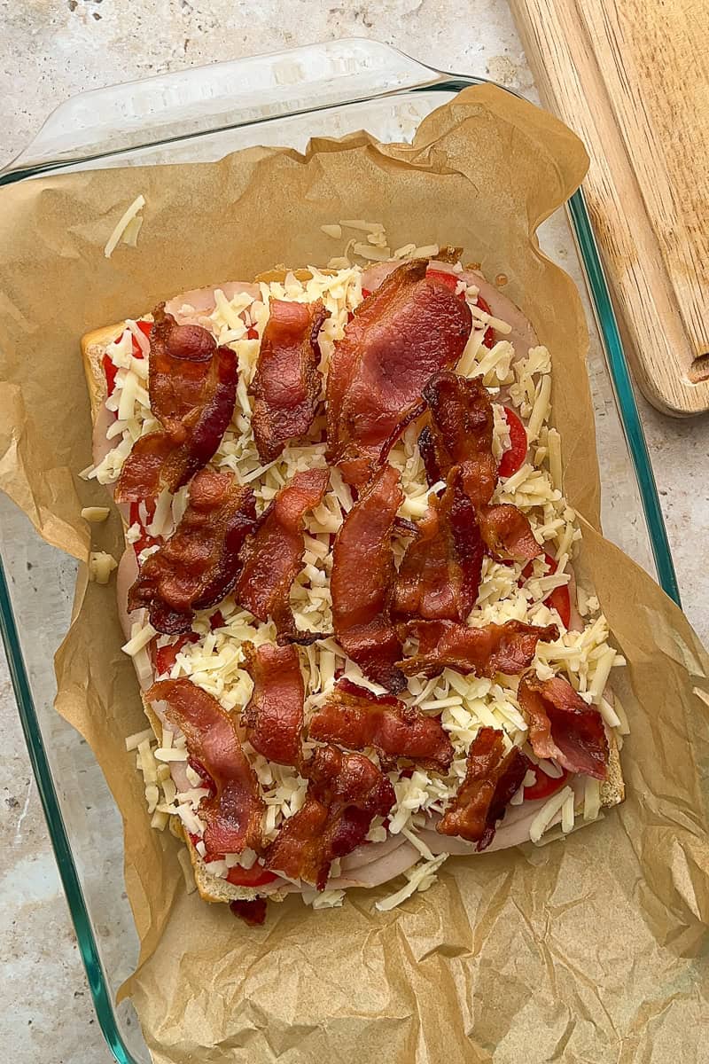 Layered hot brown sliders with turkey, bacon, tomatoes, and shredded cheese on Hawaiian rolls before baking.