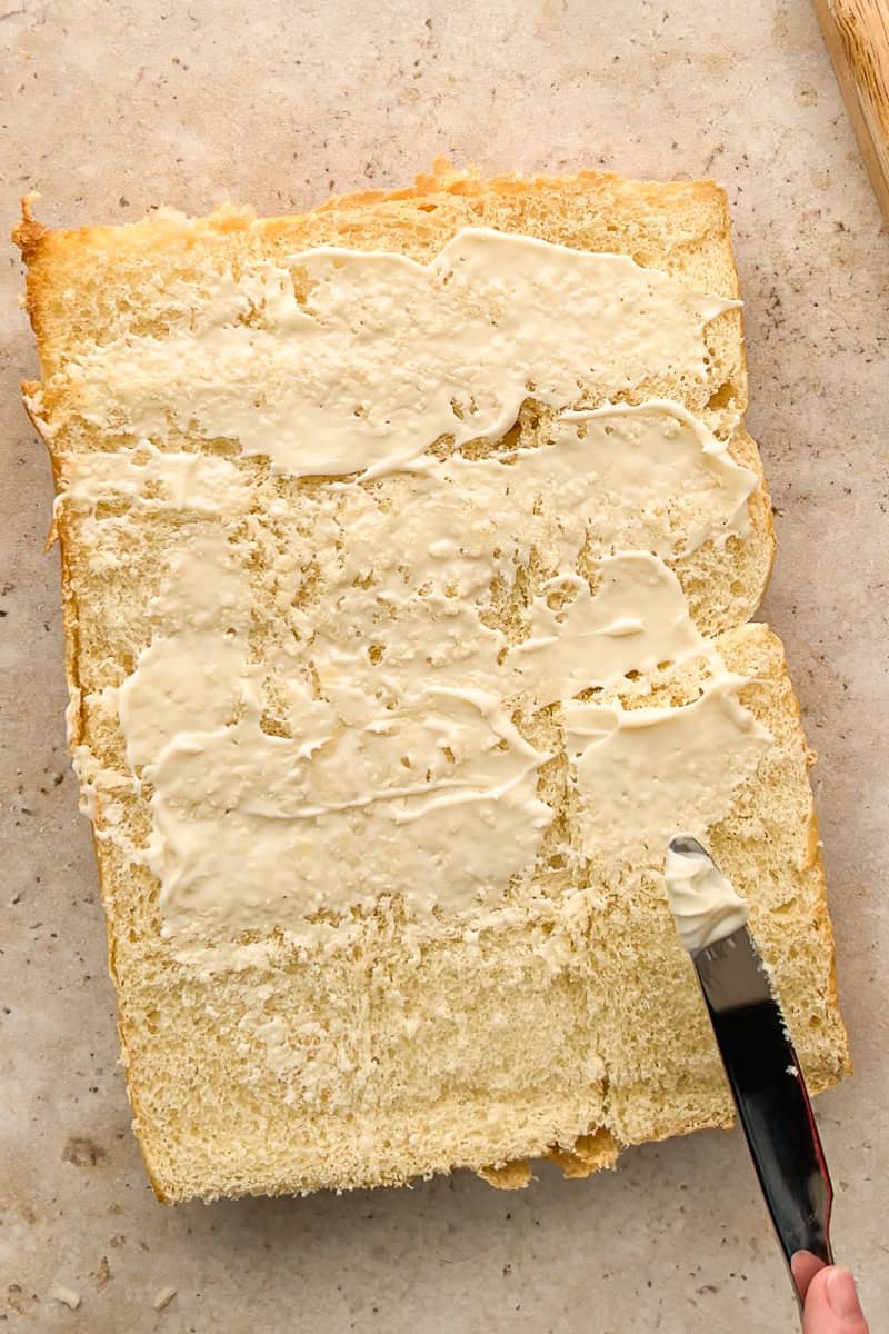 Mayonnaise being spread evenly over Hawaiian rolls as the base layer for hot brown sliders.