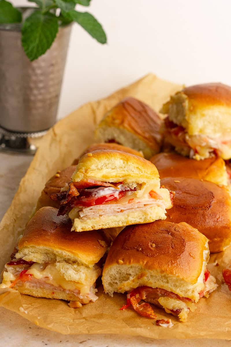 Baked hot brown sliders on Hawaiian rolls with melted cheese, turkey, bacon, and tomatoes served on parchment paper.