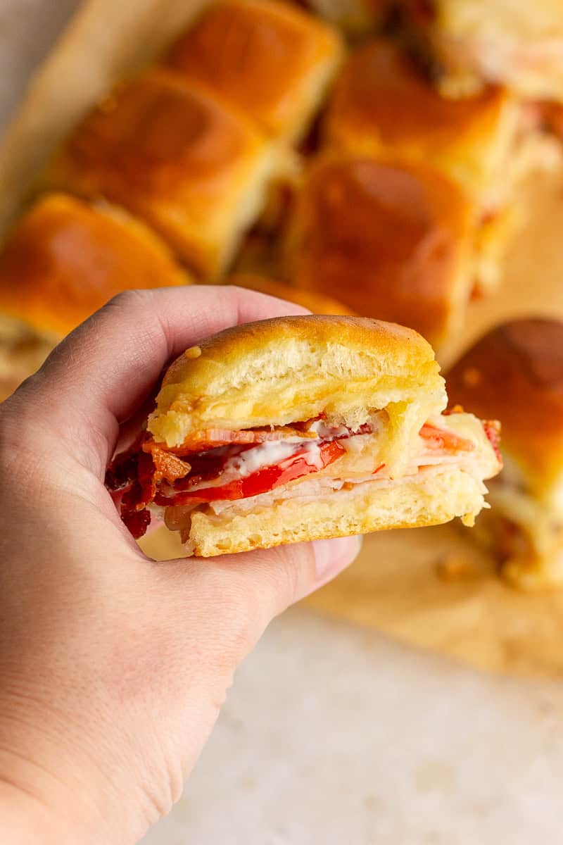 Hand holding a hot brown slider showing layers of turkey, bacon, tomato, and melted cheese on a soft roll.