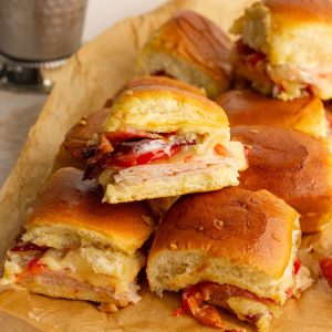 Close-up of baked hot brown sliders with melted cheese, turkey, bacon, and tomatoes on Hawaiian rolls.