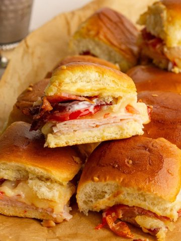 Close-up of baked hot brown sliders with melted cheese, turkey, bacon, and tomatoes on Hawaiian rolls.