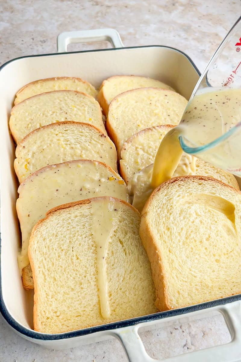 Custard mixture being poured over overlapping brioche slices in a baking dish.