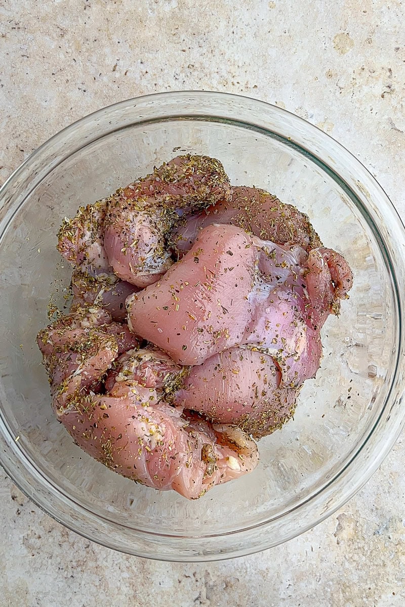 Raw chicken thighs seasoned with Italian herbs, salt, and pepper in a glass bowl before baking.