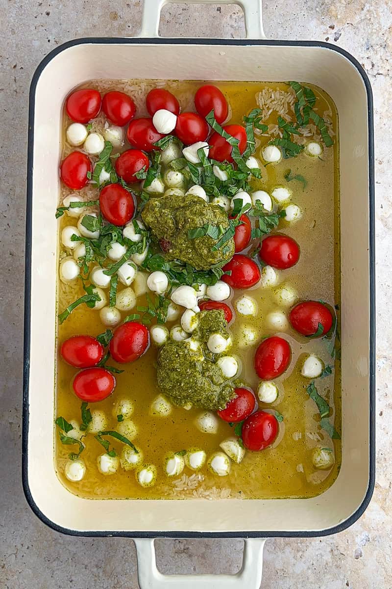 Uncooked jasmine rice with chicken broth, pesto, cherry tomatoes, mozzarella pearls, and basil in a baking dish.
