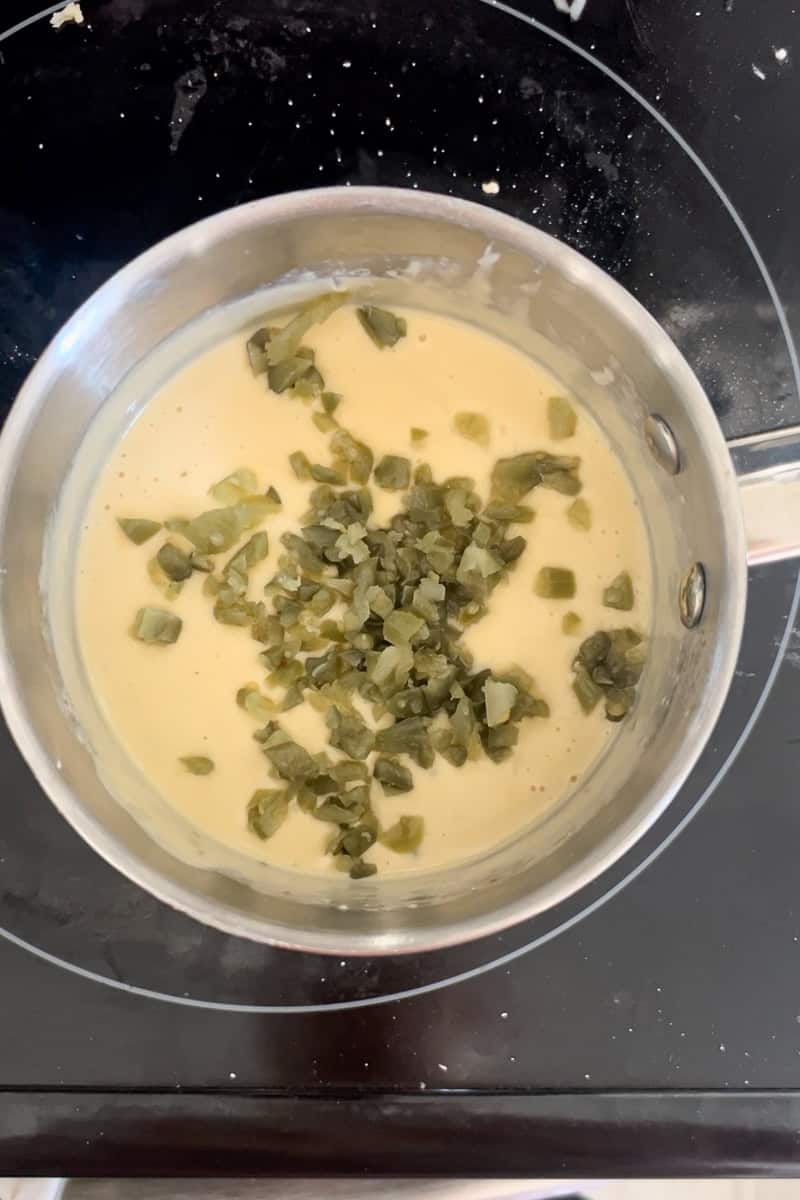 Chopped pickled jalapeños being added to a saucepan of melted queso on the stovetop.