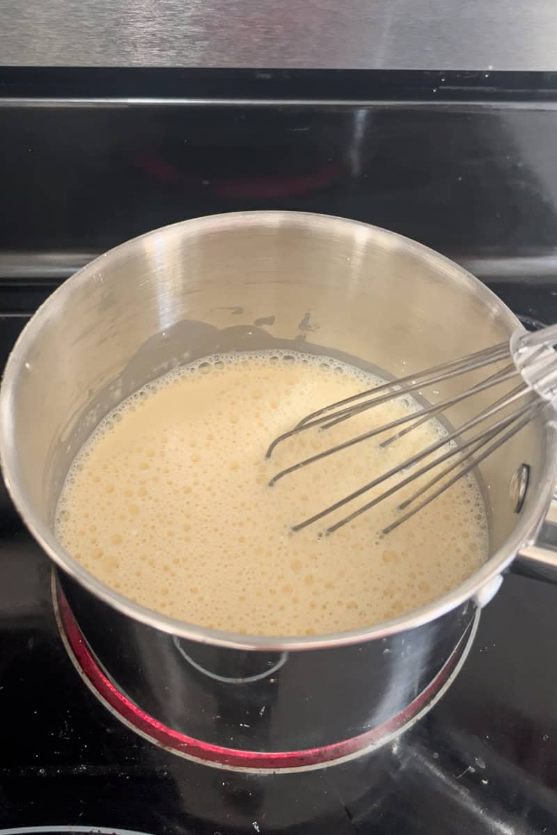 A whisk mixing evaporated milk and cornstarch in a saucepan on the stovetop.