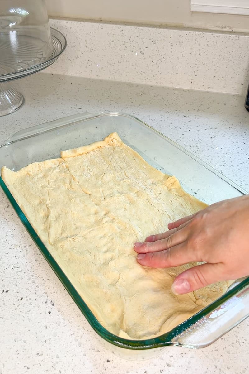 Hand pressing crescent roll dough evenly into a baking dish to create the casserole base.